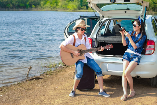 Couple of traveler sitting on hatchback of car and enjoying for view of nature near the lake during holiday.Young couple tourist have a guitar and map and baggage taking photo on vacation. 