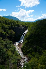 Vertical photo of Ñivinco fall in Neuquen Argentina with trees and plants in the foreground and a water stream and mountains in the background