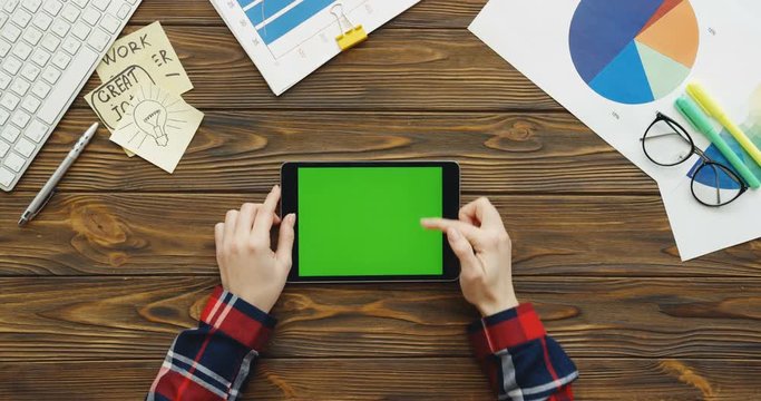 Top view on the female hands with the motley sleeves taping and scrolling on the black tablet device with a green screen horizontally on the wooden office table with office stuff, keyboard, charts and