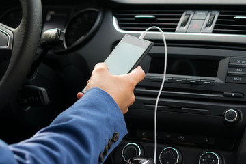 the driver of the vehicle, holds in his hand the phone connected by a white wire, to the car's music system - obrazy, fototapety, plakaty