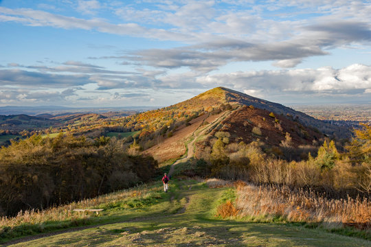 Impressive ridge of the Malvern hills, Worcestershire, England, showing a person walking on a footpath to the summit. Surrounding countryside is golden brown and orange colours of autumn and sunset