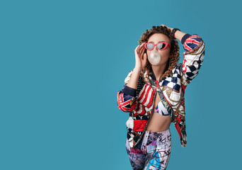 Young cute girl with curly hair in red sunglasses puffs on chewing gum on a blue background