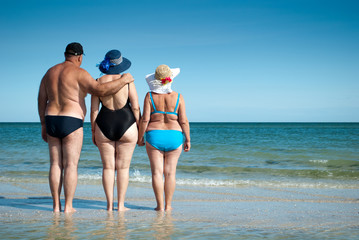 three elderly people stand on the seashore holding hands, look at the horizon resting sunbathing bathing suits swimming trunks, straw hat beach shore sand summer weekend vacation