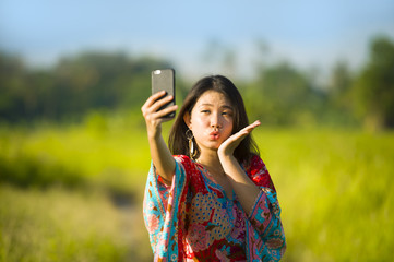 young beautiful and happy Asian Chinese tourist woman on her 20s with colorful dress taking selfie pic with mobile phone camera on tropical field