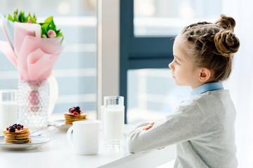 side view of child sitting at table with bouquet of flowers, glass of milk and homemade pancakes