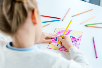 selective focus of kid drawing greeting postcard for mother with colorful pencils at table, mothers day holiday concept