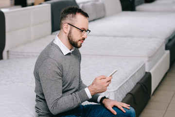 side view of male shopper with smartphone sitting on mattress in furniture shop