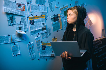 female hacker with laptop standing in front of wall with newspaper clippings
