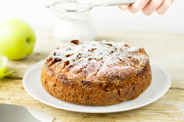 HOMEMADE RUSTIC APPLE CAKE PIE POWDERED WITH SUGAR ON A WHITE PLATE ON A WOODEN TABLE