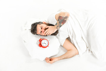 Man with yawning face lies on pillow near alarm clock.
