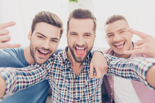Close up portrait of cool, crazy, funky, funny, attractive, stylish guys with open mouth, bristle, stubble, gesturing  rock and roll sign shooting selfie on smart phone sitting in living room