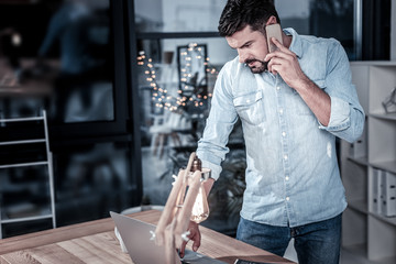 Business and evening. Concentrated occupied stylish man standing near his desktop having phone conversation and looking at his laptop.