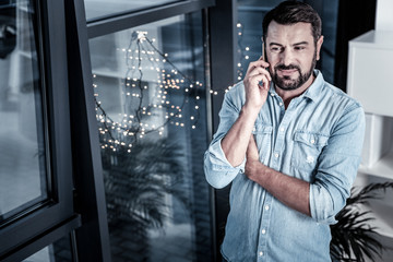 Best friends call. Pleasant handsome occupied man standing in the room near the window holding left hand under right hand and having phone conversation.