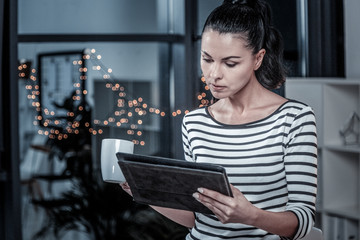 Important information. Serious cute interested woman standing in the comfortable room holding a cup and using her tablet.