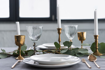 close up view of rustic table setting with wine glasses, eucalyptus, vintage cutlery, candles in candle holders and empty plates