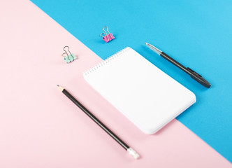Note book next to pencil, ballpoint pen and colorful clips on pink and blue background. Mockup.