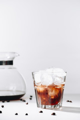 close up view of glass of cold brewed coffee, coffee maker and roasted coffee beans on grey background