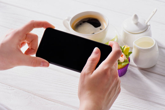 cropped shot of woman with smartphone with blank screen, cup of coffee, jag of cream and sweet muffin on wooden surface