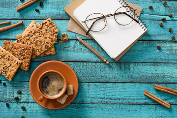 Obraz na płótnie Canvas flat lay with cup of coffee, cookies, eyeglasses, empty notebook, roasted coffee beans and cinnamon sticks around on blue wooden tabletop