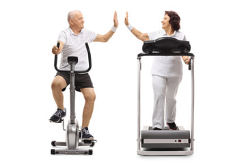 Elderly man exercising on a stationary bike and an elderly woman on a treadmill high-fiving each...
