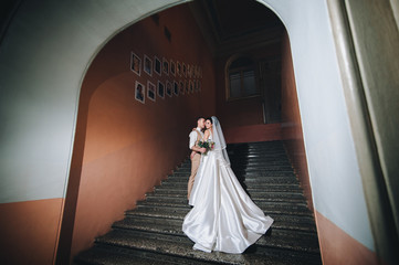 A handsome groom in a beige vest and a bride in a long dress are standing in an arch. Newlyweds on the steps.