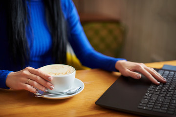 Woman working in the cafe behind a laptop, drinking coffee. The girl is typing on the laptop keyboard. Organization of the workplace