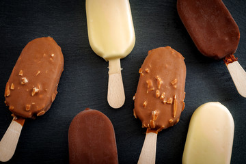 Summer desserts and cold snacks concept with many many lollies or ice cream bars on a stick in assorted flavors -  white chocolate, hazelnut and classica - on a dark black stone background