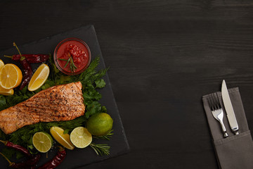 Fototapeta na wymiar top view of grilled salmon steak, pieces of lime and lemon, sauce and cutlery on black surface