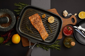 top view of arranged grilled salmon steak on wooden cutting board, sauce, ingredients and cutlery on black tabletop