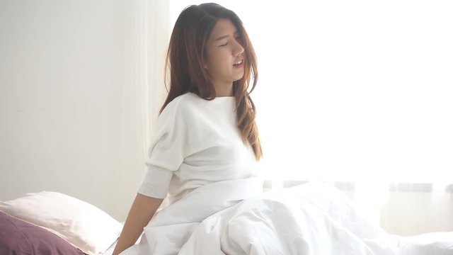 Slow motion - Happy beautiful young Asian woman waking up in morning, sitting on bed stretching in cozy bedroom looking window. Funny asian woman after wake up. Asia woman is stretching and smiling.