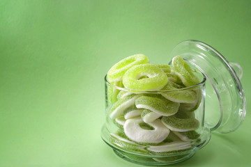 Chewy sweets and apple flavoured gummy candy concept with close up on sweet and sour green rings covered in granulated sugar in a clear glass jar isolated on a green background with copy space