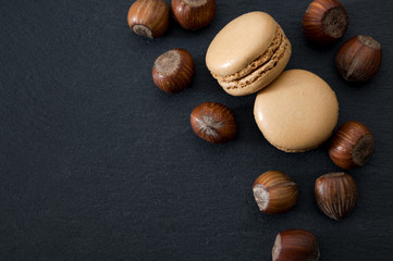French dessert, sweet meringue based confectionery and expensive sweets concept with top view of beige hazelnut flavored macaroon or macaron with a bunch of hazelnuts on dark stone with copy space