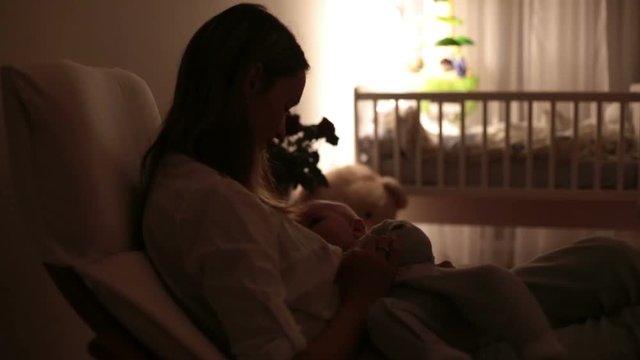 Young beautiful mother, breastfeeding her newborn baby boy at night, dim light. Mom breastfeeding infant