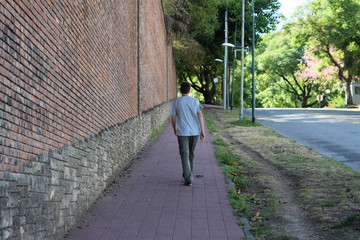 the man walks along the road along the brick wall which rises up the hill