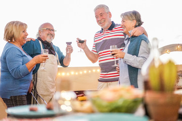 Senior friends having fun drinking wine at barbecue dinner in terrace outdoor - Mature people dining and laughing togheter - Joyful elderly lifestyle and friendship concept - Focus on left man face