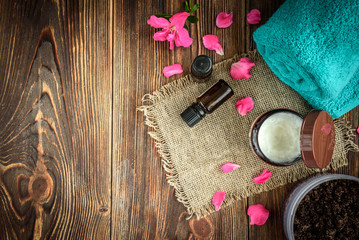 Coffee body scrub, sugar and coconut oil, essential oils, massage vacuum jars on dark wooden rustic table with pink flowers. Homemade cosmetic for peeling and spa care.