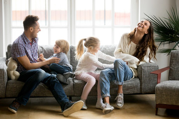 Happy parents and kids having fun tickling sitting together on sofa, cheerful couple laughing...