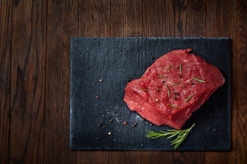 Raw meat beef steaks on black slate board with spices and rosemary over wooden background, copy space