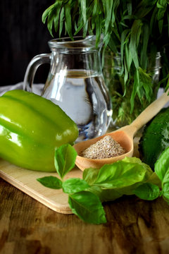 A green pepper, brans in a wooden spoon, basil, tarragon, a cucumber and water in a glass jug. Ingredients for preparing a healthy meal