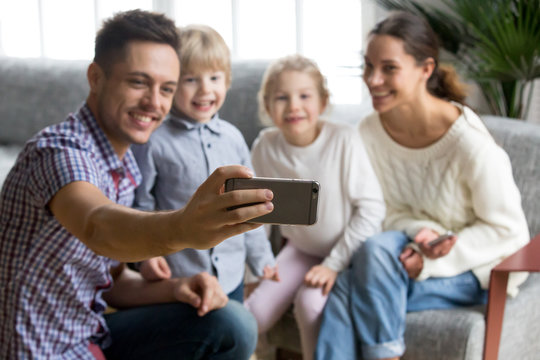 Smiling father holding smartphone taking selfie portrait of happy family with kids bonding together, husband making photo or shooting video of wife and children on mobile front camera, focus on phone