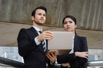 two business people in suit talking and reading information about finance news in laptop computer together standing in modern city, investment, network technology, internet and teamwork concept