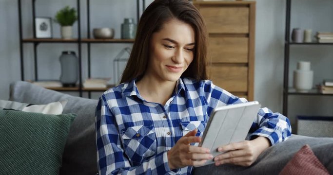 Attractive young woman in the blue plaid shirt holding a tablet computer and typing on its screen at home. Indoors