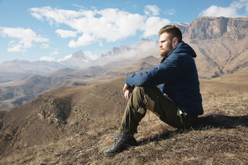 A portrait of a stylish bearded hipster sitting on a rock against the backdrop of epic rocks and contemplating into the distance thinking about life. The concept of tranquility and solitude with