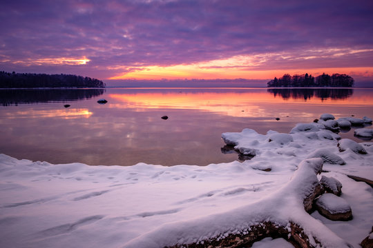 winter image of a lake at sunset with snow and stones in the foreground and an island in the background