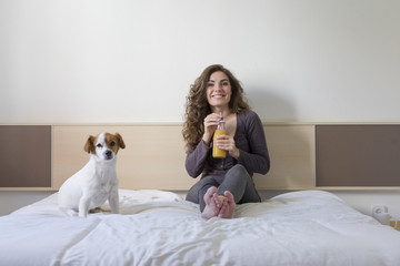 beautiful young woman sitting on bed with her cute small dog besides. Home, indoors and lifestyle. She is drinking orange juice. Breakfast at home. Happy life.