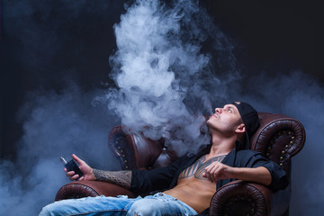 Vaper. The man with tattoos sits on a leather sofa smoke an electronic cigarette on the dark...