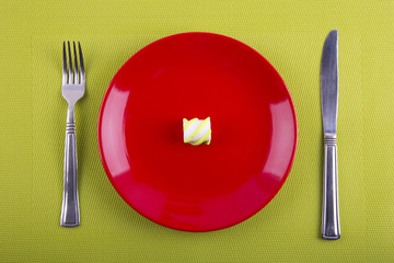 One little marshmallow on red round plate with knife and fork on green background
