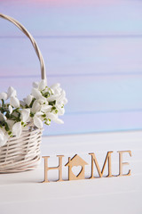 Wooden inscription "Home" and bouquet of tender snowdrops in wicker basket on light blue background.