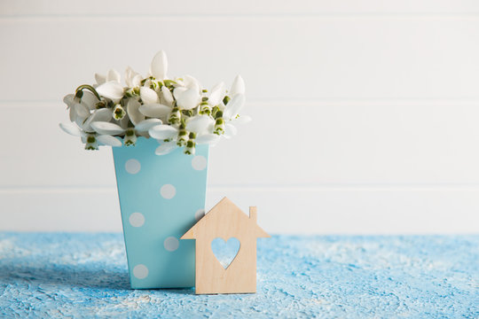 Wooden house with hole in form of heart with spring snowdrop flowers in blue paper box on white background