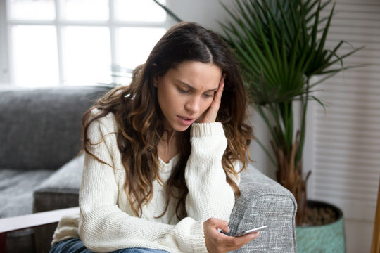 Sad girl feeling upset reading bad news in mobile message on smartphone at home, depressed frustrated young woman holds phone scared of threatening suffering from cyberbullying being bullied via cell